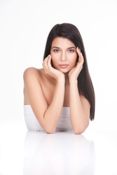 a portrait of a young woman, posing on a white background. she has both her elbows resting on a white table. both her hands are touching her face delicately,