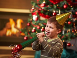 Little boy laughing on new year's eve, wearing shiny hat and blowing horn, looking at camera, christmas tree in background.?