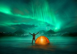 A man camping in wild northern mountains with an illuminated tent viewing a spectacular green northern lights aurora display. Photo composition.
