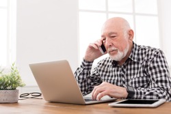 Senior man talking on phone and using laptop, sitting at table at home. Modern technology, communication concept