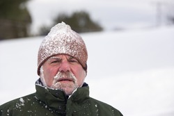 A man wearing a beanie cap and covered with snow is looking at the camera with an unhappy expression. Horizontal shot.