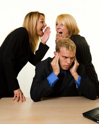One business man with hands covering hears sitting at desk with two woman coworkers talking behind his back