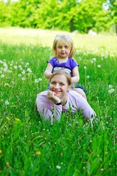 Daughter sits on mother on a grass outdoors smiling