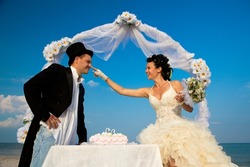 Bride and Groom Under Archway on Beach with wedding cake. Bride putting bit of cake on groom's nose