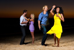 People (two couples) on the beach having a party and lots of fun in the sunset, they are wearing smart casual clothes and dance