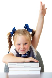 Blonde happy schoolgirl in blue dress and matching tie rises her hand up, while sitting at school desk with pile of books, isolated on white