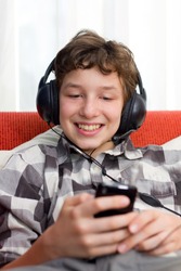 A preteen boy having fun as he listens to his mp3 player through big headphones on his head. He's seated on an orange couch with white pillows and he's wearing a checkered dress shirt.