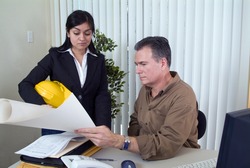 A woman holding a hardhat looking at what appears to be a set of plans held by a man.