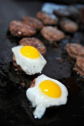 Several mini Kobe beef burgers cooking on a grill. Two are topped with a fresh fried quail egg.