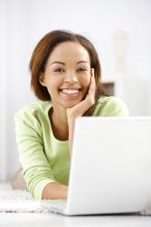Portrait of laughing ethnic girl lying on floor, using laptop computer, looking at camera happily.?