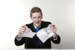 man in a suit sat at a desk ripping a piece of paper up with the words debit card printed on it and looking happy doing so.