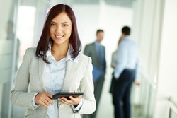 Business woman standing in foreground with a tablet in her hands, her co-workers discussing business matters in the background, tilt up