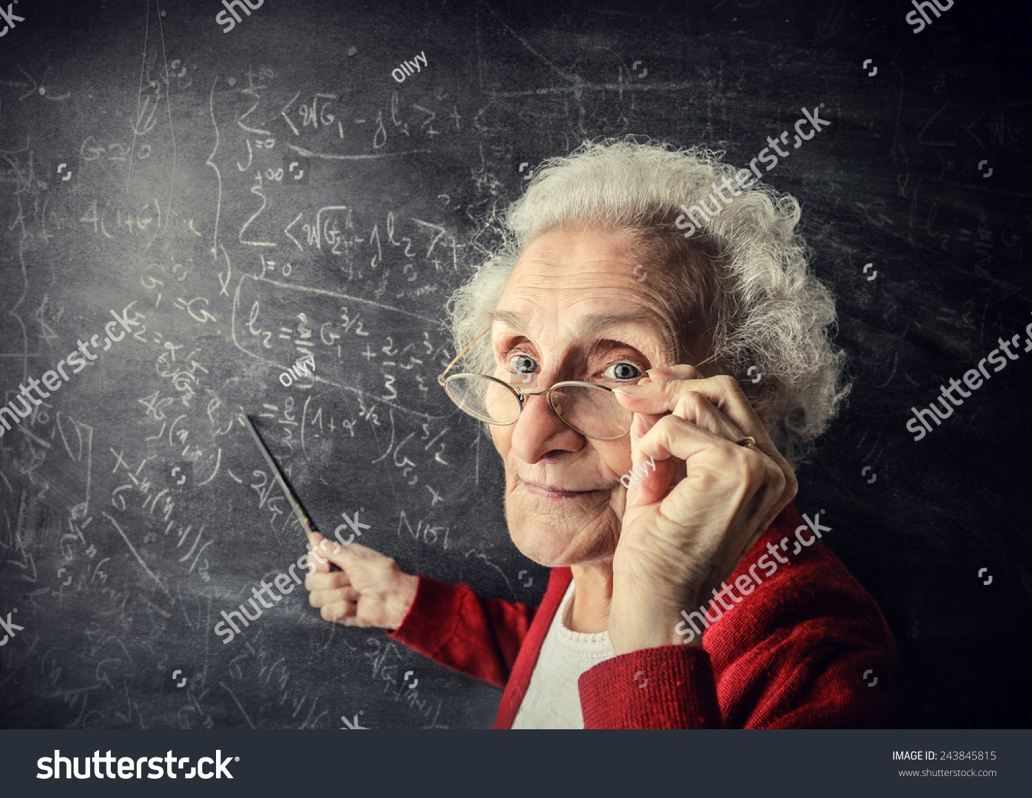  An elderly woman with white hair and glasses is standing in front of a blackboard full of mathematical equations, holding a stick and pointing at the board while looking at the camera.