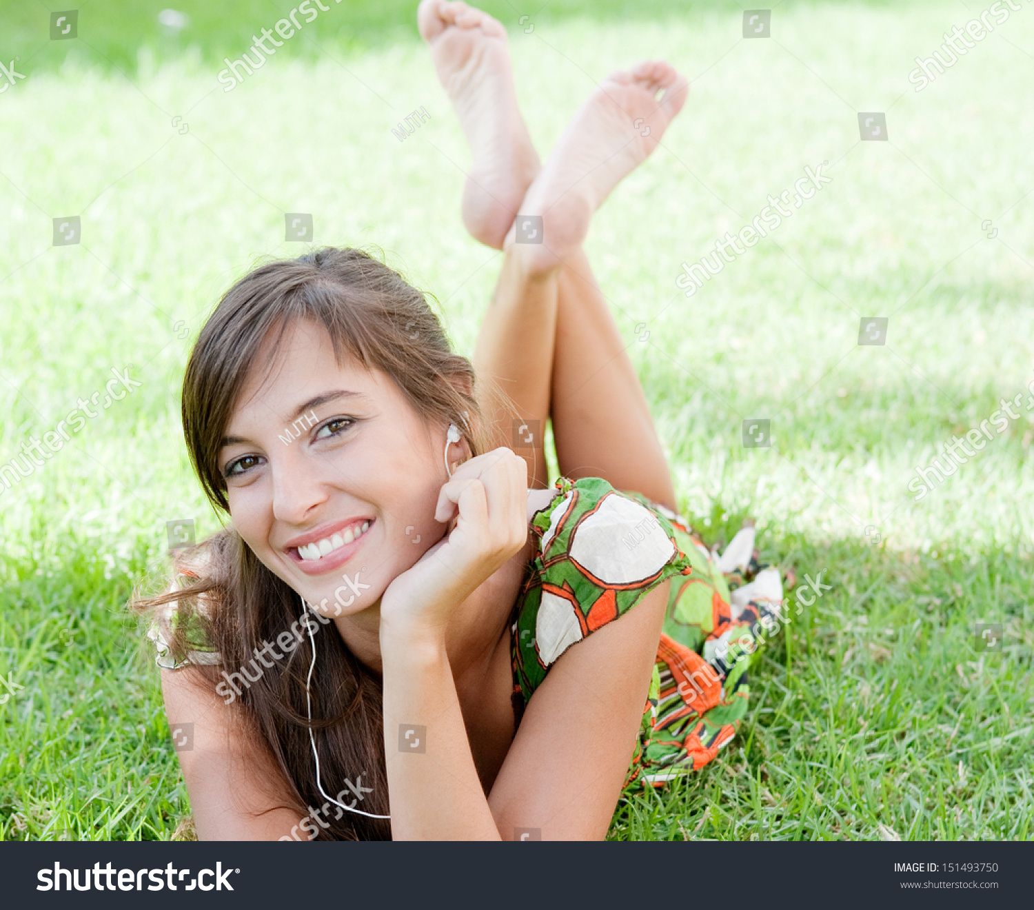 Attractive Young Woman Laying Down On Fresh Green Grass In A Park With Her Feet Up And Listening