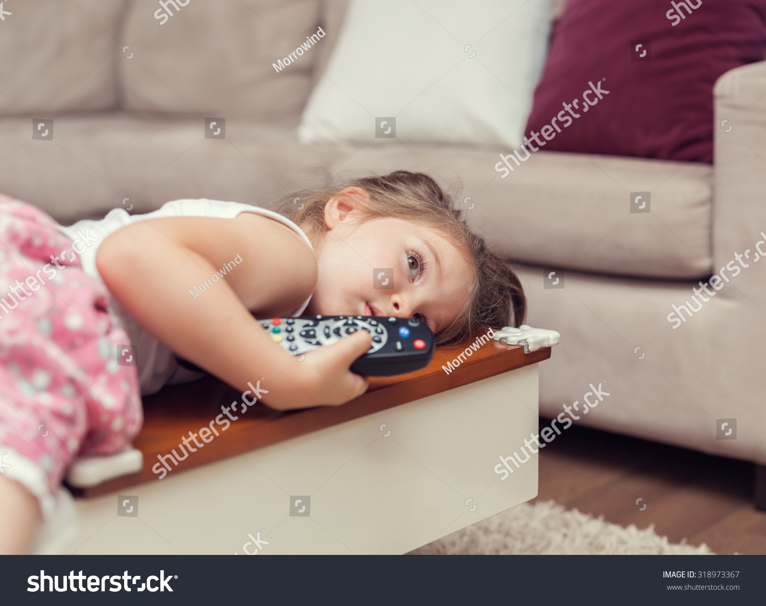 Cute Little Girl With Remote Control Lying On Coffee Table And Watching