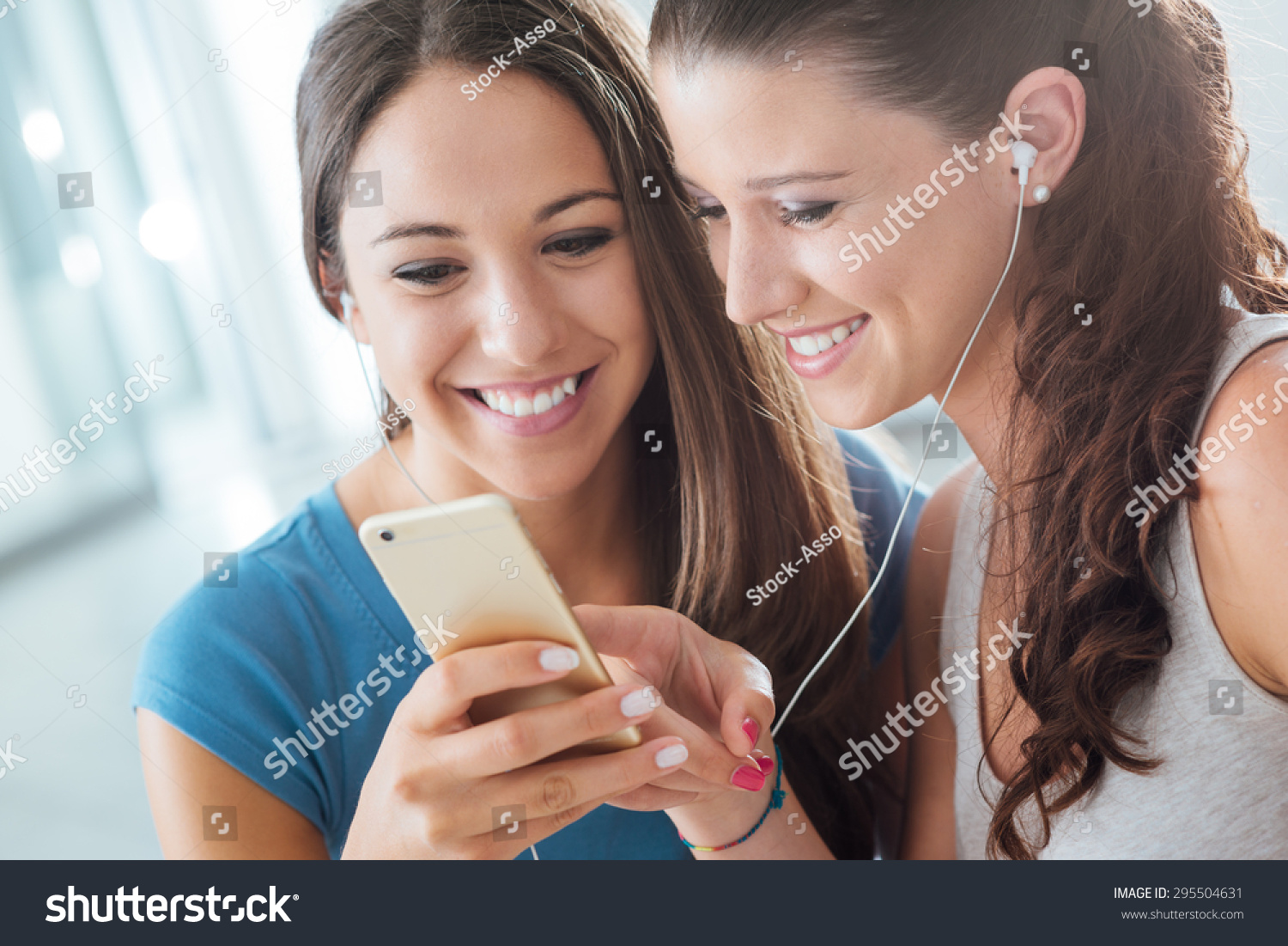 Pretty smiling young girls sharing earphones and listening to music using a smart phone Pretty smiling young girls sharing earphones and listening to music using a smart phone