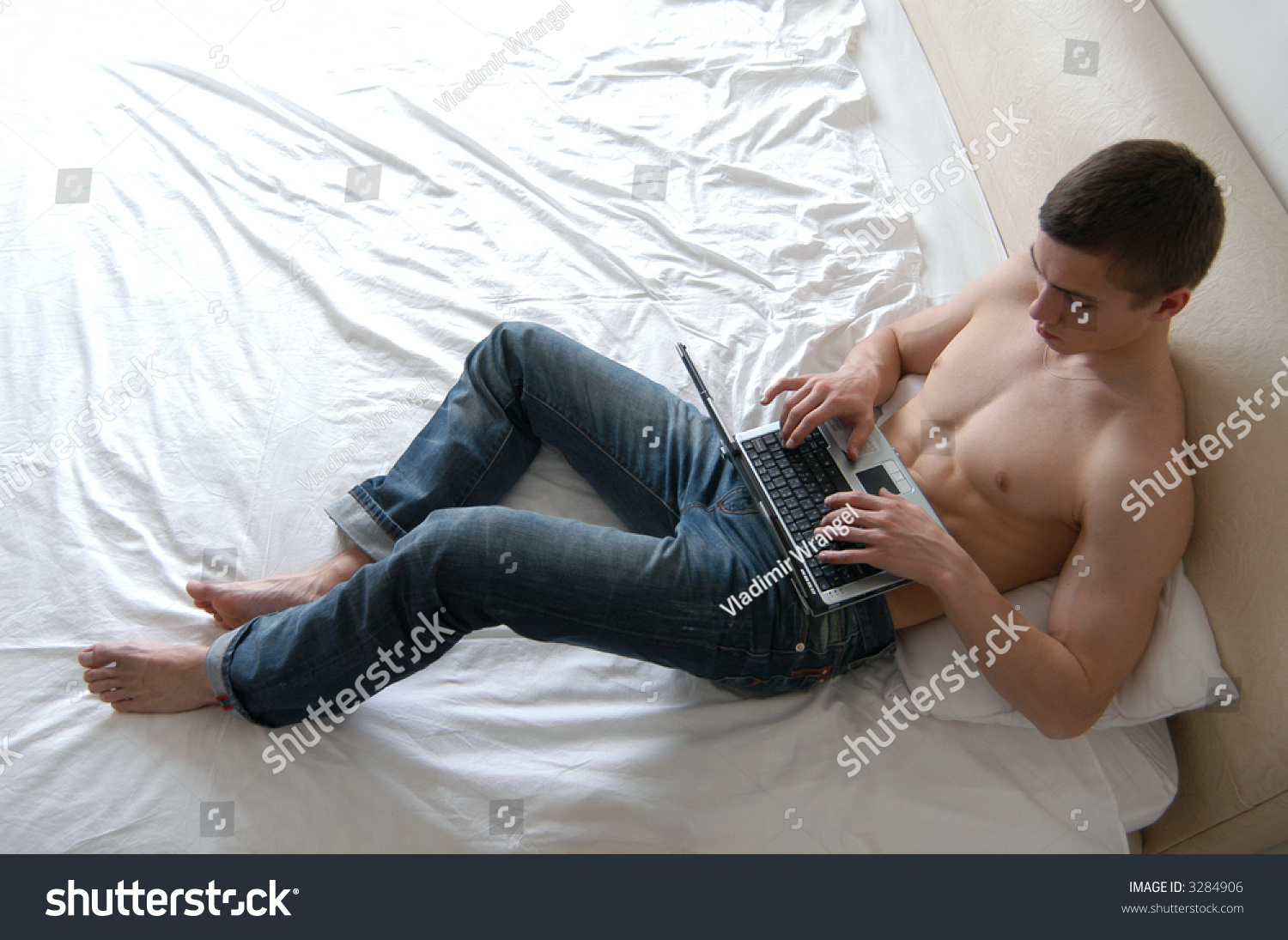 Shirtless Young Man Working On A Laptop Lying On The Bed Stock Photo