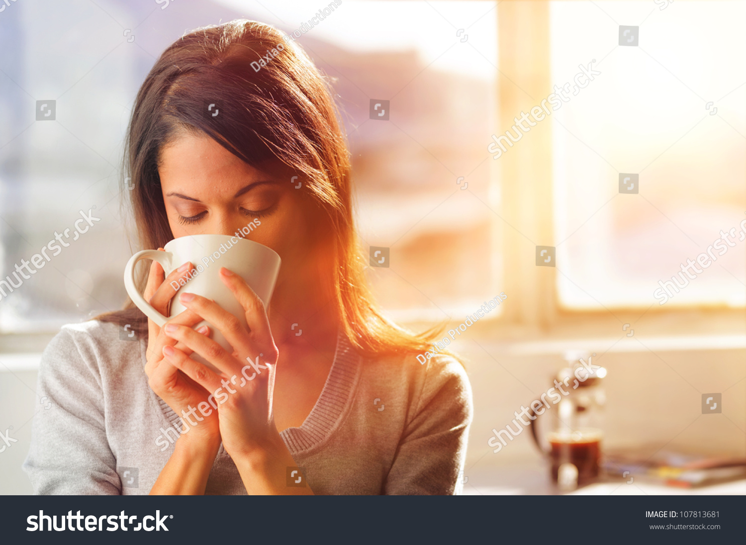 Woman Drinking Coffee At Home With Sunrise Streaming In Through Window 