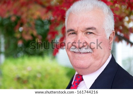 White hair senior businessman looking at you around fall time under the tree with red leaves behind him