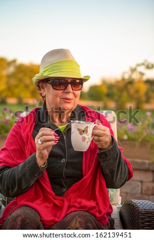 Senior woman with a straw green hat, sitting at the patio and showing her hot tea, perhaps she wants to stay warm