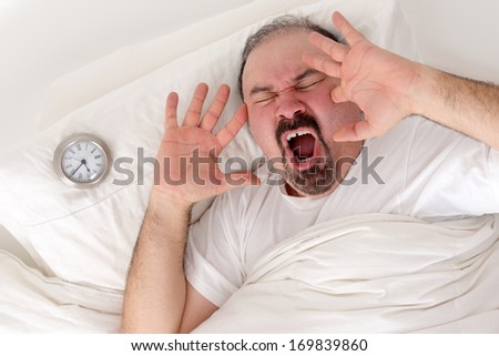 Middle-aged bearded man yawning loudly resting in bed with his alarm clock on the pillow alongside him as he tries to wake after a sleepless night