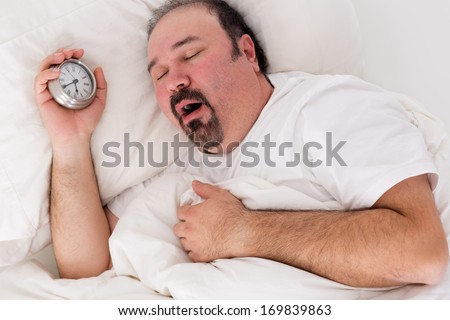 Lethargic tired man lying in bed yawning as he struggles to wake up unmotivated to start the new day and content to rather continue lying in bed as he holds his alarm clock in his hand