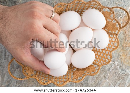 Hand of a man wearing a wedding ring taking a traditional Easter egg from an ornamental woven wicker Easter basket filled with fresh white eggs