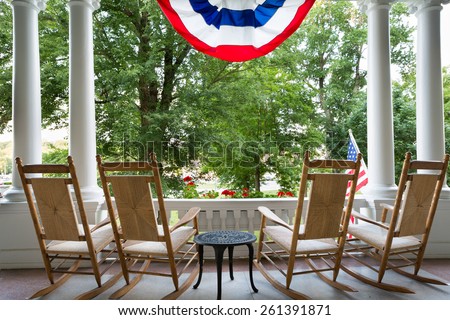 Four vacant wooden rocking chairs lined up on a patio overlooking a lush garden below a draped American flag symbolizing 4th July commemorating Independence Day
