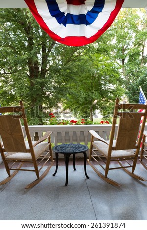 Two Wooden Rocking Chairs with Small Table at the Terrace with Conceptual American Flag Fronting Tall Green Trees at the Garden