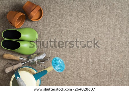 Side border of gardening tools and equipment including green garden shoes, flowerpots, hand tools and a watering can arranged in a line, overhead view with copyspace on a beige textile