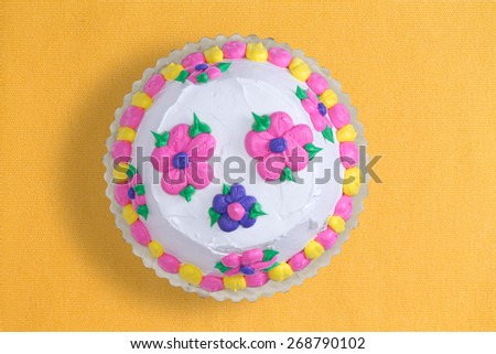 Top view of a decorative iced homemade cake with colorful pink and blue flowers surrounded with a yellow and pink alternating border, served uncut on a yellow background with copy space