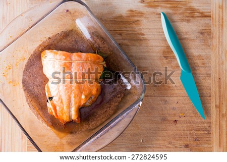 Fresh oven-baked salmon fresh from the oven in a glass oven dish alongside a sharp blue kitchen knife on a wooden kitchen counter ready to be served, view from above