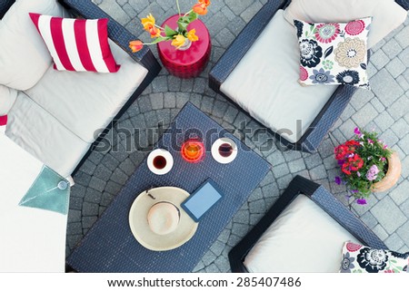 Relaxing on a brick patio by candlelight in the evening with two cups of tea and a straw sunhat on a table surrounded by deep seating comfortable armchairs and flowers, overhead view