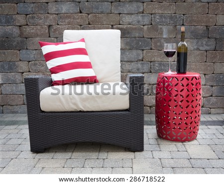 Comfortable Dark Wicker Patio Chair Outfitted with Plush Cushions Beside Modern Red Table with Bottle and Glass of Red Wine on Outdoor Stone Affluent Patio
