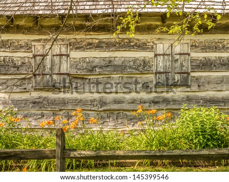 Two rustic wood shutters on a log cabin with trees, flowers and wood fence in front of the cabin.
