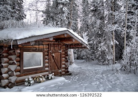 A log cabin frosted over in sub zero temperatures, with a trail leading behind it down to the river.