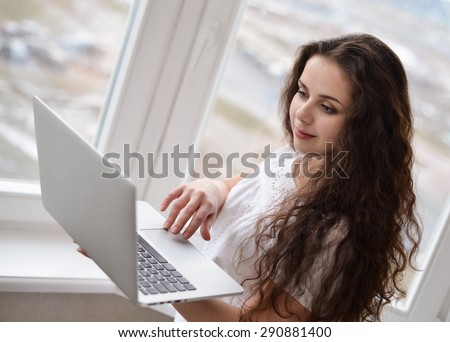 A young woman holding a laptop,standing near window.