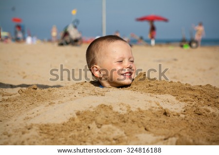 Boy buried in sand on the sea behind head smiling. Little baby boy hiding himself in sand on the beach. Only head is visible. Smiling face and happy.