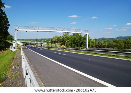 Electronic toll gate above an empty highway in a wooded landscape. Bridge and forested mountains in the background. White clouds in the blue sky.