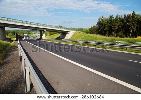 White truck entering under a concrete bridge over a highway in a wooded landscape. Noise protection wall. White clouds in the blue sky.