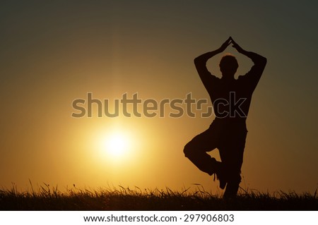 Silhouette of a man in a position Vrikshasana at exercising yoga on a grassy horizon at sunset.
