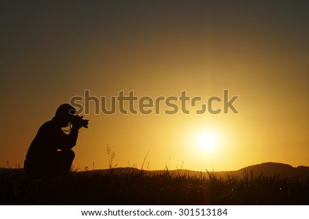 Silhouette photographer kneeling on a grassy horizon at sunset. Wooded mountains in the background.