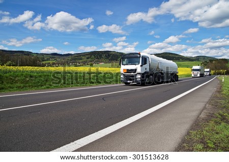 White tanker and trucks driving along the asphalt road around the yellow flowering rapeseed field in countryside. Wooded mountains in the background. Blue sky with white clouds.