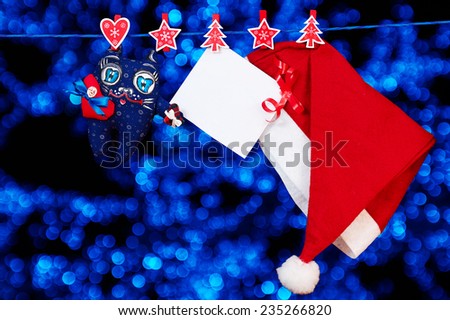 Christmas decoration santa hat and a cat toy and empty sheet of paper with a red ribbon hanging on rope over blue blurred lights background