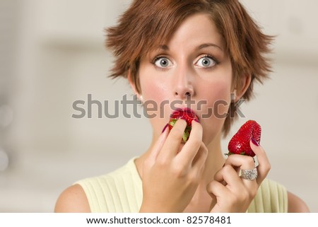 Pretty Wide-eyed Red Haired Woman Biting Strawberry in Her Kitchen.
