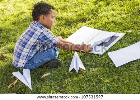 Mixed Race Boy Learning How to Fold Paper Airplanes Outdoors on the Grass.