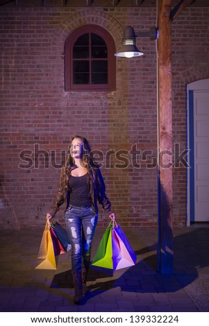 Excited Pretty Mixed Race Young Adult Woman Walking in the Evening Holding Shopping Bags with Brick Wall Background.