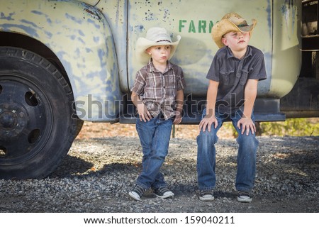 Two Young Boys Wearing Cowboy Hats Leaning Against an Antique Truck in a Rustic Country Setting.