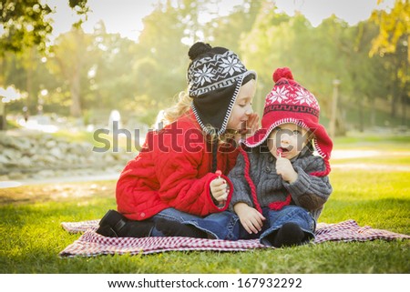 Little Girl Whispers A Secret to Her Baby Brother Wearing Winter Coats and Hats Sitting Outdoors at the Park.