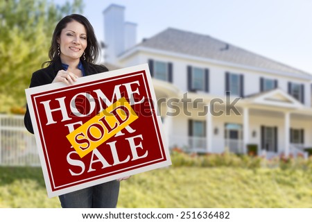 Smiling Hispanic Woman Holding Sold Home For Sale Sign In Front of Beautiful House.